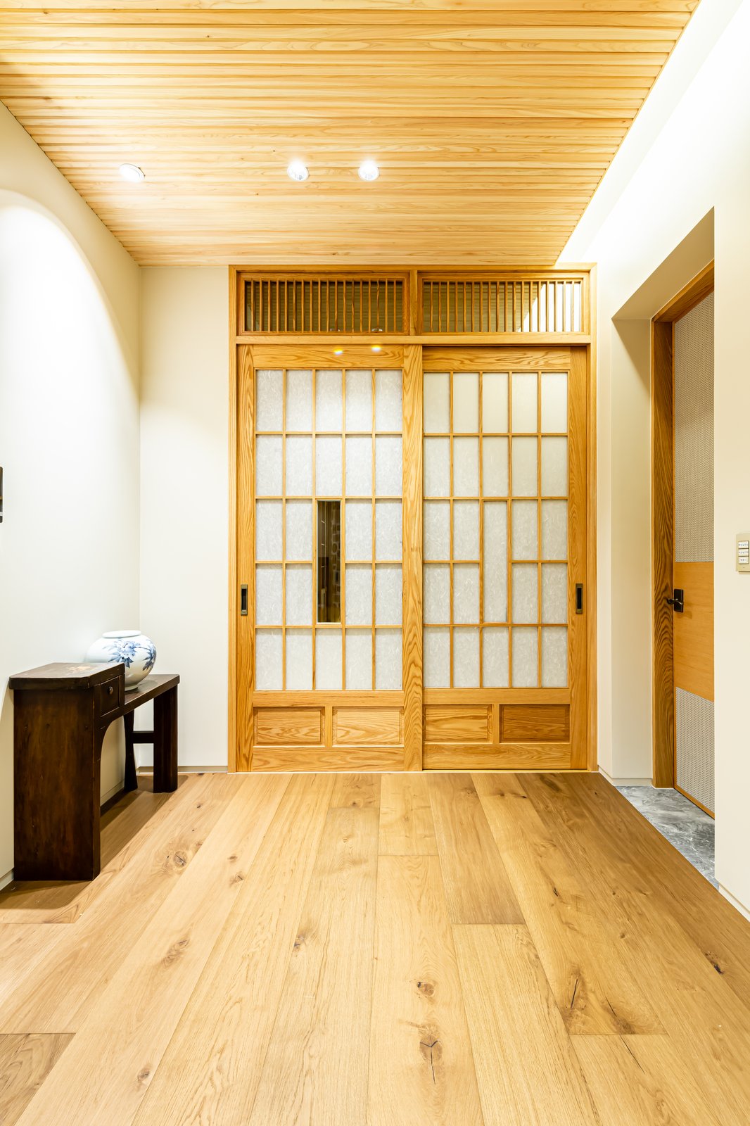 Entryway with paneled shoji sliding doors beneath a wood-plank ceiling, dark side table with a blue-and-white vase