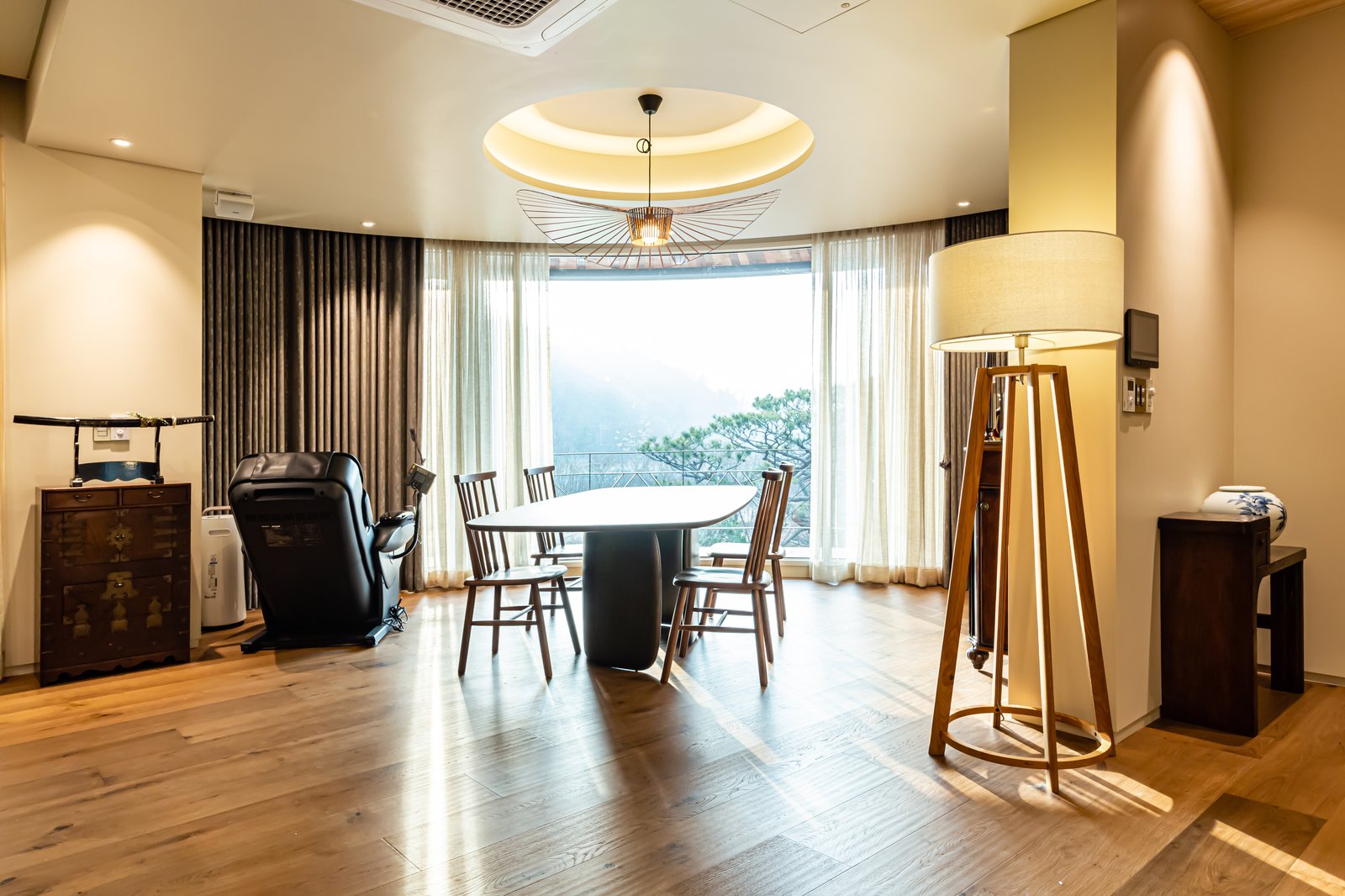 Round dining table and yellow sofa beneath a circular skylight and pendant light, floor-to-ceiling curtained windows with mountain view