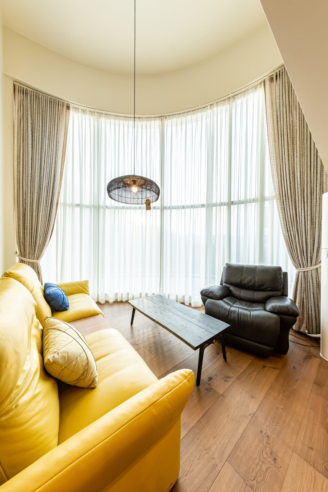 Living room corner with a mustard-yellow leather sofa and black recliner facing tall curved curtained windows