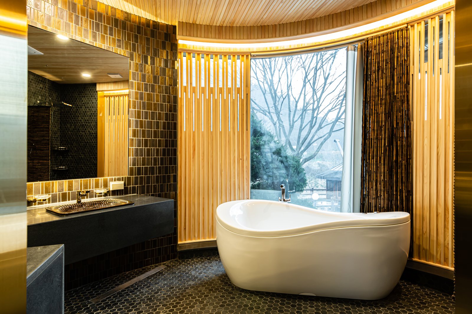 Bathroom with gold mosaic tile walls, a freestanding tub, and a wood-slatted window wall overlooking bare trees