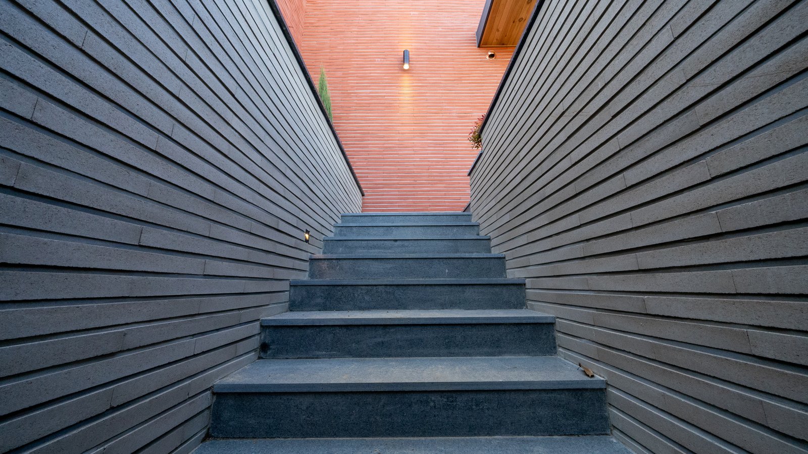 View up a stone stairway between wood-slat walls toward a red brick wall with a sconce